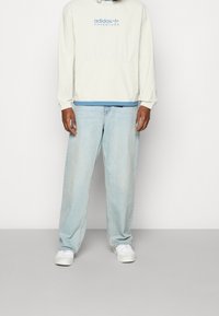 Ljusbeige hoodie med blå logotyp, kombinerad med ljusblå raka jeans och vita sneakers. Tyget verkar mjukt och avslappnat.