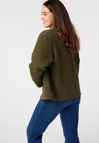 Olijfgroene gewatteerde jas met een getextureerd bladenpatroon, featuring een losse pasvorm en design met zijzakken, gecombineerd met blauwe jeans.