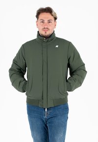 Giacca a vento verde con colletto alto, zip frontale, tasche laterali e polsini a costine. Presenta un piccolo logo sul lato sinistro.