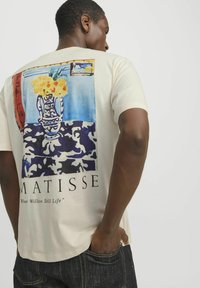 Cremfarbener Baumwoll-T-Shirt mit einem bunten Druck eines Vasen mit Blumen, Kunstwerk von Matisse, mit fettem Text und einem strukturierten Muster am unteren Rand.