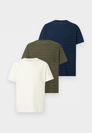 Mężczyzna z kręconymi włosami, ubrany w granatowy t-shirt z krótkim rękawem, białe spodnie i brązowe buty, stoi z rękami w kieszeniach na białym tle.