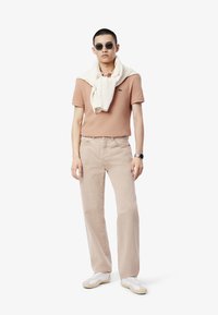 Beige polo paita, villanein valkoinen neule hartioilla, vaaleanbeige housut, valkoiset lenkkarit ja tummat aurinkolasit; minimalistinen muotoilu.