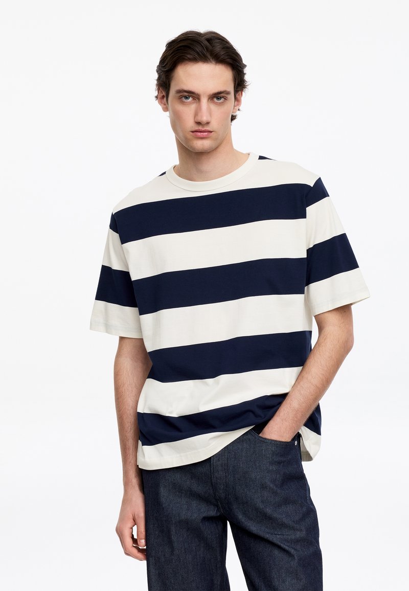 Jeune homme aux cheveux foncés portant un t-shirt à manches courtes avec de larges rayures horizontales bleu marine et blanc et un jean foncé, une main dans la poche.