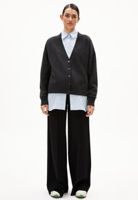 Donkergrijze gebreide cardigan met een V-hals, knoopsluiting en geribde manchetten, gelaagd over een lichtblauw shirt en brede zwarte broeken.