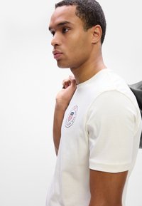 Hvid kortærmet t-shirt med rund halsudskæring og et cirkulært logo med røde og blå detaljer på venstre bryst. Glat tekstur.