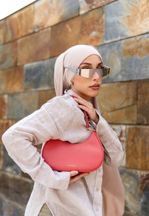 Vrouw met beige hijab en oversized reflecterende zonnebril houdt koraalroze Coccinelle handtas vast tegen een achtergrond van een stenen muur.