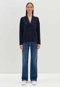Navy blazer with lapels and two front pockets, paired with blue flared jeans and white sneakers. The fabric is smooth with a structured design.