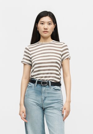 Γυναίκα με μακριά σκουρόχρωμα μαλλιά που φοράει καφέ και λευκή ριγέ κοντομάνικη μπλούζα, μπαγιάτικη μέσα σε ψηλόμεσο ανοιχτό μπλε τζιν με μαύρη ζώνη.