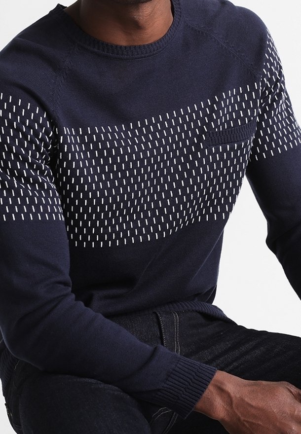 Homme portant un pull bleu marine avec des motifs en tirets blancs verticaux sur la poitrine et les bras supérieurs, assorti à un jean foncé.