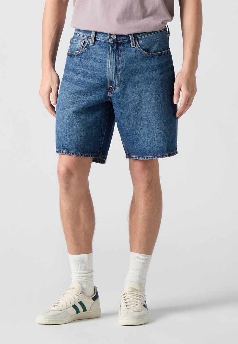 Mand iført blå denimshorts i midterlængde, hvide sokker og hvide sneakers med mørkegrønne striber på en lys baggrund.