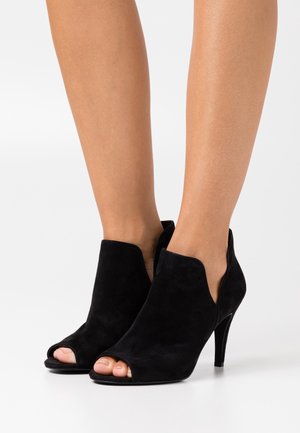Botines de ante negros de punta abierta con tacón que presentan un diseño con cortes y una textura suave, mostrando un estilo minimalista y una silueta elegante.