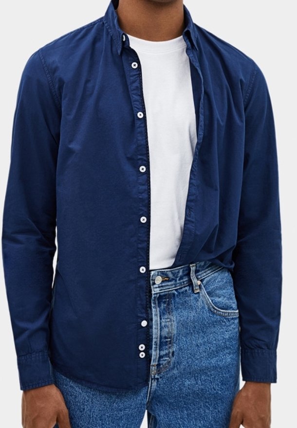 Granatowa koszula zapinana na guziki z kołnierzykiem, długimi rękawami i białymi guzikami, noszona na białej koszulce, zestawiona z jasnoniebieskimi dżinsami.