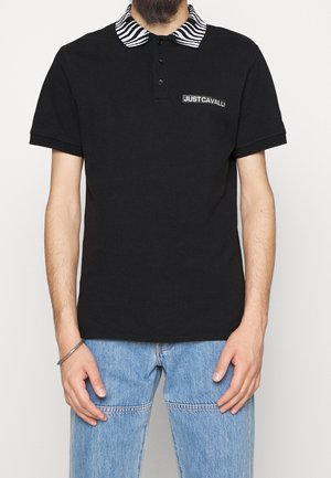 Mężczyzna w czarnej koszulce polo z kołnierzykiem w paski zebry oraz logo "JUST CAVALLI", w połączeniu z jasnoniebieskimi dżinsami.