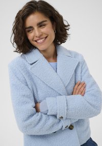 Femme avec des cheveux bruns courts et bouclés souriant, portant un manteau texturé bleu clair et croisant les bras devant un fond blanc uni.