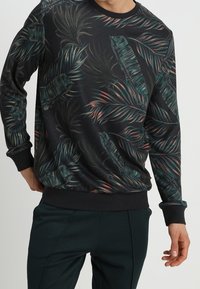 Uomo che indossa una felpa nera con un motivo di foglie tropicali verdi e rosa, e pantaloni scuri, in piedi con una mano in tasca.