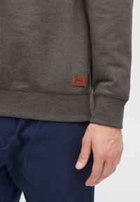 Mørk grå sweatshirt med ribbet nederkant og manchetter, med et brunt lædermærke syet på nederste venstre side. Blå bukser synlige.
