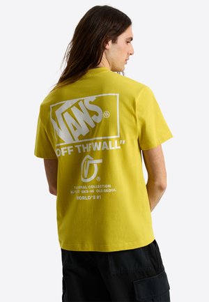 Mężczyzna z długimi włosami noszący żółtą koszulkę Vans z dużym białym logo "Vans Off The Wall" i czarne krótkie spodnie typu cargo, zwrócony tyłem.