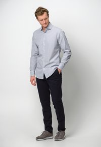 Gestreept overhemd in blauw en wit, lange mouwen, gecombineerd met donkere broeken en grijze sneakers, staande op een neutrale achtergrond.