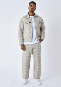 Chaqueta de mezclilla con estampado y pantalones a juego en color beige claro con diseños geométricos. Lleva una camiseta blanca debajo y zapatillas blancas.