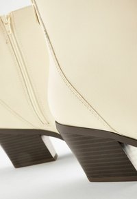 Bottines en cuir crème avec des talons empilés marron foncé et des fermetures éclair sur les côtés, sur un fond blanc.