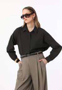 Black button-up shirt with loose fit, paired with high-waisted beige trousers, accented by a black belt. Model wears oversized sunglasses.