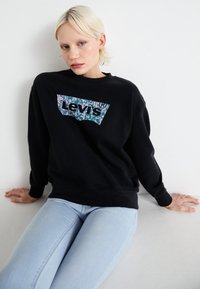 Čierna mikina so mnohofarebným kvetinovým logom Levi’s. Bavlnený materiál, voľný strih, rebrované manžety, čistý pozadí, nosená s svetlomodrými džínami.