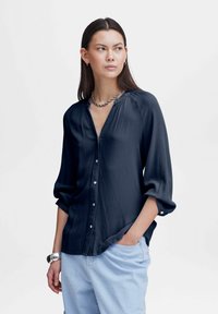 Donkerblauwe blouse met lange mouwen en een V-hals, met textuur, knoopsluiting en opgerolde mouwen. Draagt met lichtblauwe broek.