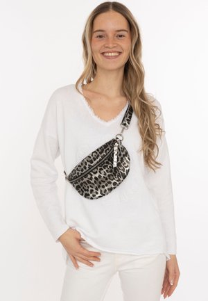Lächelnde Frau mit langem, welligem Haar, die einen weißen Pullover und eine weiße Hose trägt, dazu eine silberne und schwarze Crossbody-Tasche mit Leopardenmuster.