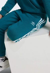 Pantalones deportivos en color azul verdoso, confeccionados en mezcla de algodón, con diseño de tres franjas blancas y logotipo en la parte inferior. Textura suave, ajustados en los puños.