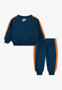 Set od dukserice i hlača u mornarsko plavoj boji s narančastim prugama. Izrađeno od mekanog materijala, s rebrastim manžetama i bijelim Nike logotipom na leđima.