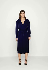 Vestido envolvente azul-marinho de mangas longas com um decote em V profundo e um laço na cintura, feito de um tecido suave. Usado com sapatos de salto agulha pretos e com tiras.