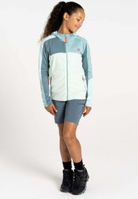 Hellblaue, mit Reißverschluss versehene Kapuzenjacke mit einem dunkleren blauen Panel, kombiniert mit grauen Shorts. Hat eine weiche Textur und kontrastierende orangefarbene Akzente. Schwarze Wanderstiefel.