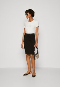 Camiseta blanca con el logo "GUESS", falda lápiz negra ajustada, tacones de slingback negros y un bolso de mano con patrón en blanco y negro.
