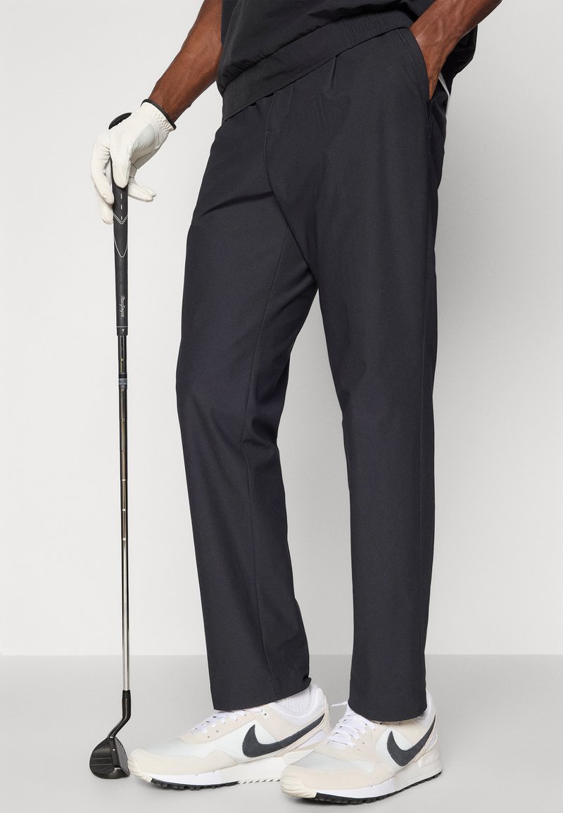 Pantaloni da golf grigio scuro con vestibilità rilassata, dotati di tasche e una texture liscia, abbinati a scarpe sportive bianche e a una mazza da golf.