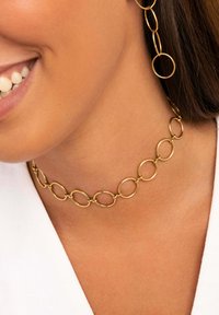 Choker de metal dorado con formas ovaladas unidas y bordes suaves, acompañado de pendientes circulares a juego, usados sobre un fondo claro.