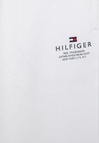 Tela blanca con bordado en negro que dice "HILFIGER" y un pequeño logo rectangular en rojo, blanco y azul por encima del texto.