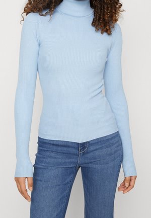 Femme portant un pull col roulé côtelé bleu clair à manches longues et un jean bleu, debout devant un fond clair uni.