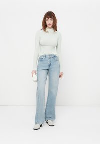 Un col roulé à côtes vert clair, un jean bleu clair taille haute, un sac à main texturé blanc, des chaussures à talons noires et blanches, le modèle se tient contre un fond blanc.