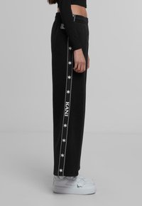 Črne trenirke z belim vzorcem zvezd in besedilom "KANI" ob strani, z elastičnim pasom. V kombinaciji z belimi supergami.