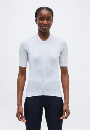 Mujer con un maillot de ciclismo de manga corta azul claro con cremallera frontal y pantalones cortos de ciclismo negros acolchados, de pie frente a un fondo liso.