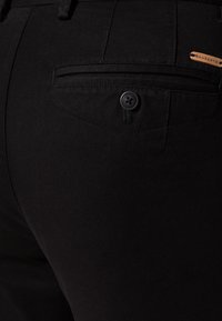 Czarne spodnie wykonane z teksturowanej tkaniny, z małą tylną kieszenią z guzikiem oraz metką marki w pobliżu pasa.