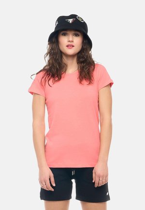 Giovane donna con capelli mossi che indossa una t-shirt corallo, pantaloncini neri e un cappello a secchiello nero con toppe, in piedi davanti a uno sfondo semplice.