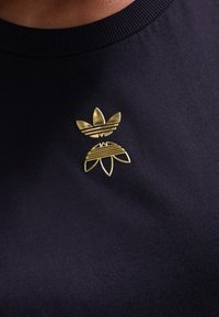 Marinfärgad T-shirt i tyg med en guldfärgad metall-logotyp som har en trekantsdesign ovanför en stiliserad blomform, båda med linjemönster.