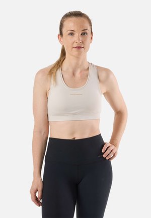 Donna con pelle chiara e lentiggini, indossa un top corto beige e leggings neri a vita alta, in piedi con una mano sul fianco davanti a uno sfondo bianco.