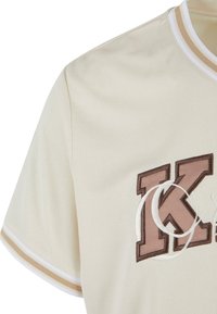 Lyse beige sportsjersey med struktureret stof, kontrasterende hvide og beige striber på kraven, samt et markant broderet brunt 'K' logo.