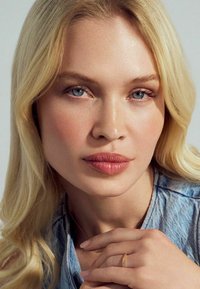 Modell med långt, vågigt blont hår och blå ögon, iklädd en denimskjorta och en delikat guldring på sitt finger, med mjuk, naturlig smink.