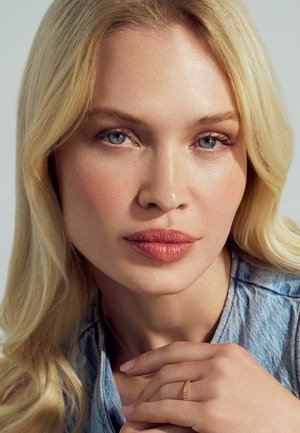 Model med langt, bølget blond hår og blå øjne, iført en denimskjorte og en delikat guldring på fingeren, med blød, naturlig makeup.