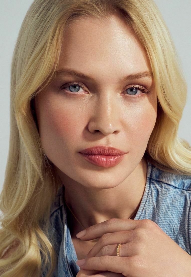 Modell med långt, vågigt blont hår och blå ögon, iklädd en denimskjorta och en delikat guldring på sitt finger, med mjuk, naturlig smink.