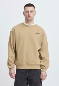 Felpa beige con vestibilità rilassata, collo a girocollo ribassato e spalle cadenti. Presenta una piccola grafica nera sul lato sinistro del petto.