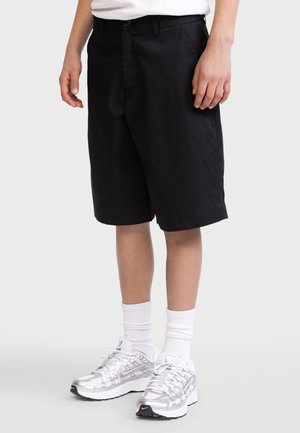 Person iført sorte knælange shorts, hvide crew-sokker og hvide og sølvfarvede sportssko stående på en ensfarvet baggrund.